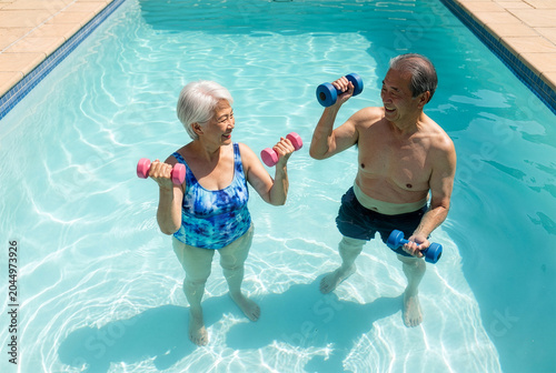 Happy active Asian senior couple exercising with dumbbells in a swimming pool on a sunny day.