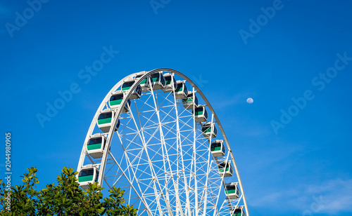 A giant ferris wheel with the moon rising on the side in the blue sky.