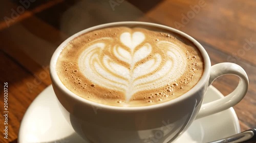 A white ceramic cup filled with a hot latte featuring elegant leaf latte art, resting on a white saucer with a metal spoon on a wooden table under natural warm sunlight.