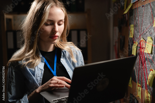 Young female detective using laptop near board with evidence or case investigation and criminal lead. crime work at night