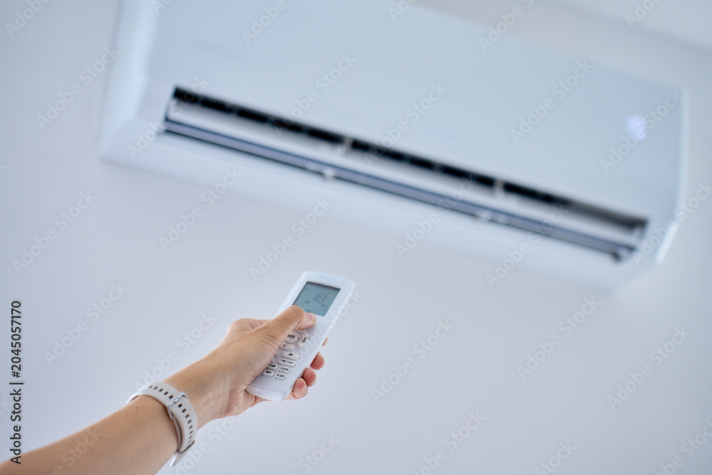 Obraz premium Hand adjusts a wall air conditioner with a remote control in a bright room. Concept of cooling, temperature setting and climate control.