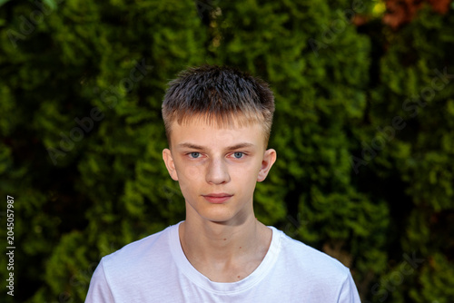 Close portrait of a serious teen boy looking at the camera with a calm expression, dark green thuja hedge in background