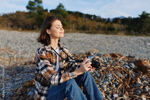Fotografia Female photographer sitting on a rocky shore with a camera, basking in sun and breeze