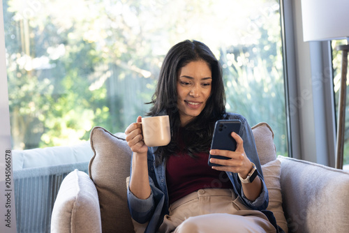 African American woman sitting on cushioned sofa by large window holding smartphone and ceramic mug