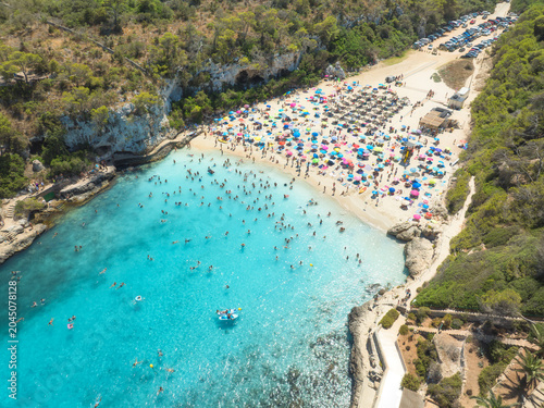 Schilderij op canvas Aerial view of colorful umbrellas on crowded sandy beach, swimmers in blue sea, green trees on sunny summer day