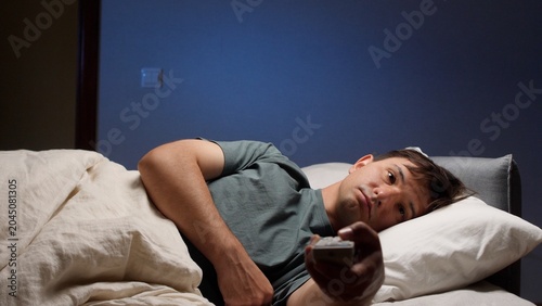 Obraz na plátně Man lying awake in dark bedroom late at night, clutching tv remote and staring a