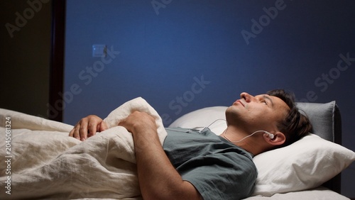 Konstfotografi Man lying on a comfortable bed in a dark bedroom, listening to an audio podcast