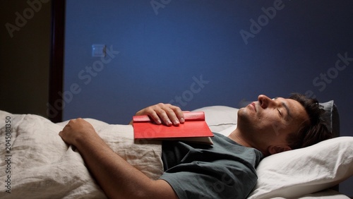 Fotografie Man sleeping peacefully on a comfortable bed in a dim bedroom, red book on his c