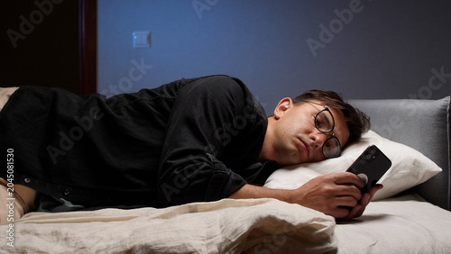 Slika na platnu Young man wearing optical glasses lying in bed late at night scrolling a smartph