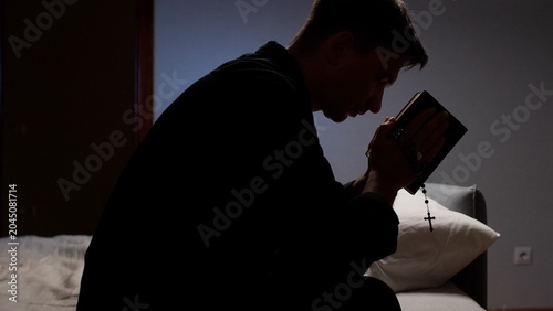 Фотография Man holding a rosary and holy bible, sitting in a dark room, deeply engaged in p