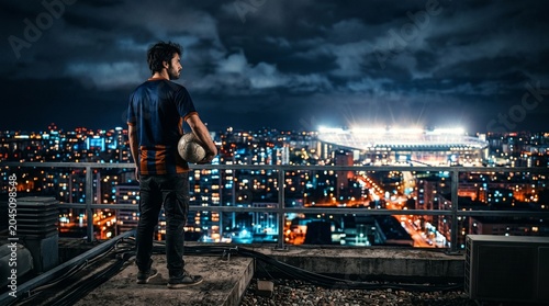 Solo soccer player standing on high rooftop overlooking glowing city stadium at night.
