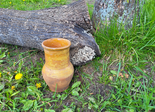 A brown clay jug stands next to a gray log on a green lawn in the garden