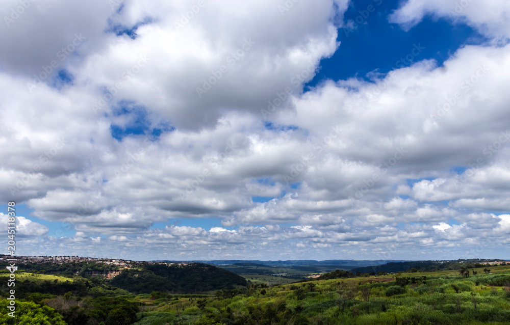 Fototapeta premium Expansive blue sky with white cumulus clouds over a vibrant green rural landscape in the Brazilian countryside on a sunny day.