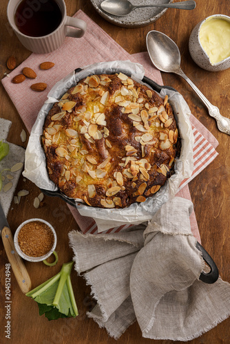 Homemade rhubarb pudding cake with flaked almonds in baking pan. Flat lay food photography of sweet seasonal dessert on rustic wooden table background. Cozy afternoon tea concept