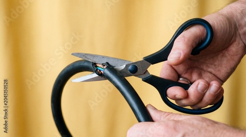 Hands inappropriately use standard scissors to sever a thick black electrical cable. This conveys the danger and folly of using incorrect tools for hazardous electrical jobs.