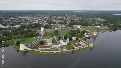 Aerial view captures ancient fortress on water's edge. Stone walls encircle buildings, red roofs stand out. Green fields stretch beyond, towns fade in distance. No people, just calm
