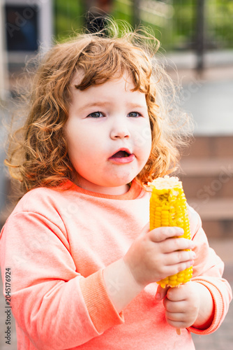 Toddler girl eating boiled yellow corn cob outdoors. Cute ginger kid with curly hair holding healthy snack. Childhood lifestyle, organic nutrition, vegetable diet and summer food concept.