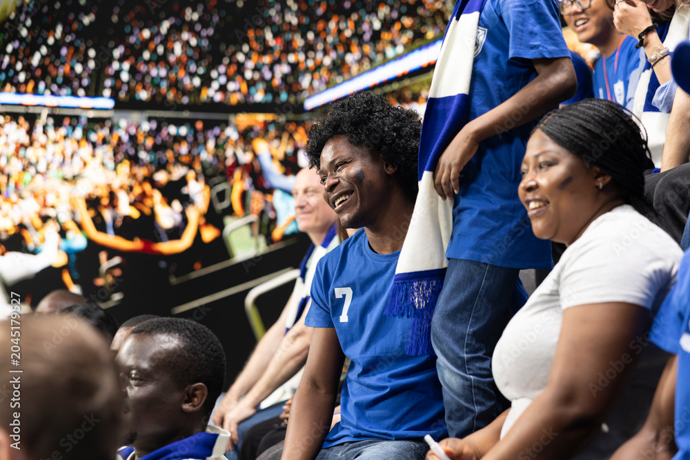Fototapeta premium Diverse blue team football supporters chanting in packed stadium stands during soccer match, fan scarves and excited crowd atmosphere. Father, mother and son sharing family fun with laughter.