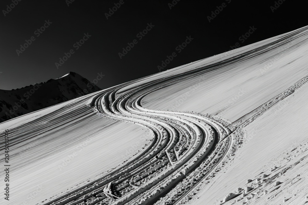 Obraz premium Black and white photo shows ski tracks on a snowy mountain slope. A dark sky looms above a distant peak. Fresh snow covers the landscape with winding trails. This scene suggests winter sport activity.