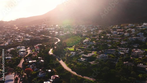 Aerial view of Cape Town hillside near Table Mountain. A drone sweeps a winding road past upscale homes and pools at golden hour, with backlight and lens flare.