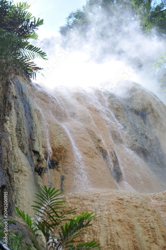 A waterfall with steam rising from it. The steam is white and the water is brown. The waterfall is surrounded by trees and plants