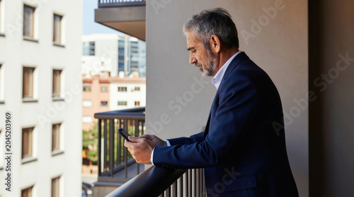 Mature man in suit stands on balcony holding smartphone. He checks messages or reads news while looking over cityscape. Casual business on terrace during morning.