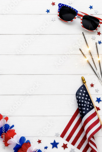Flat lay of an American flag, patriotic sunglasses, and sparklers on white wooden background with copy space for Fourth of July.