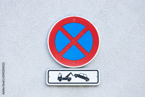 No parking sign with red circle and blue cross symbol, accompanied by towing warning sign depicting a tow truck and car, mounted on a gray wall