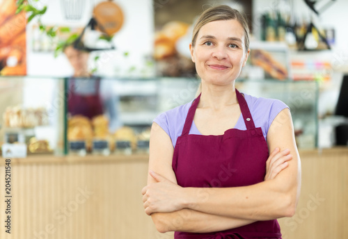 Tableau sur toile Positive young waitress wearing pinafore standing in cozy cafeteria ready to gre