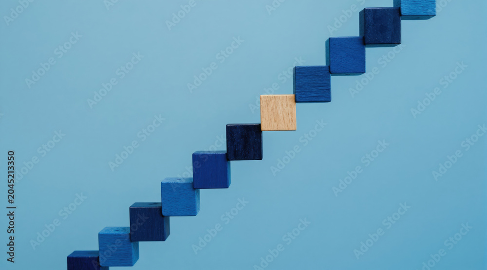 Fototapeta premium Wooden blocks form a step ladder with different blue shades. One block differs in color and texture, suggesting progress or uniqueness. Clean blue background highlights abstract concept.