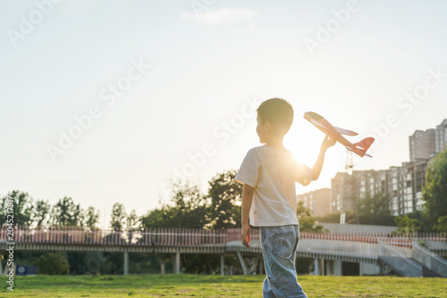 Bilde på lerret A young Asian boy holds a toy airplane while standing on a grassy field at sunset, enjoying outdoor playtime in a city park
