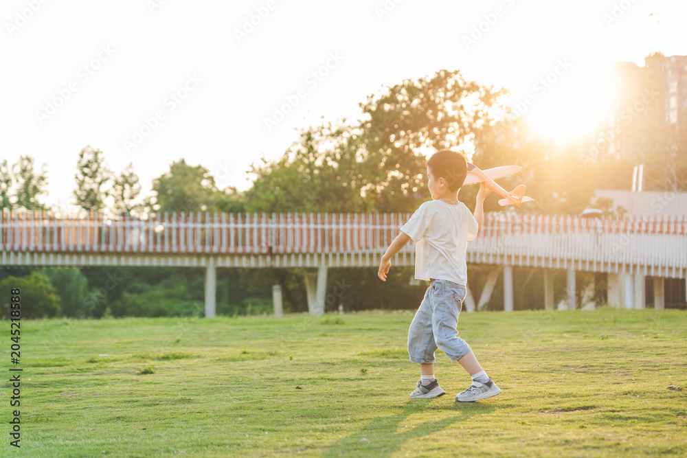 Obraz premium A young Asian boy plays with a toy airplane on a lush green lawn during golden hour sunset, with a pedestrian bridge and trees in the background.