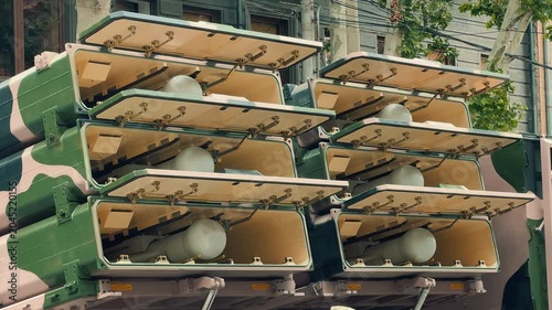 A detailed close look at a drone inside its launcher transitioning to a wide shot of multiple camouflage military trucks parked on a city street during an exhibition.
