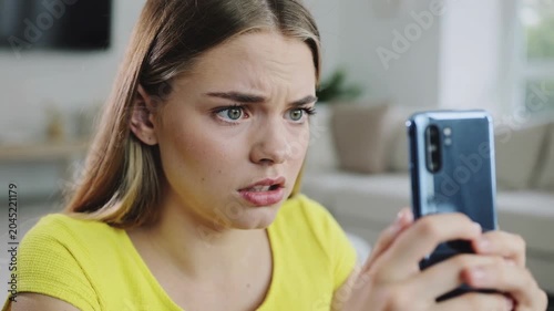 Young woman in yellow shirt looking at smartphone screen with shocked facial expression indoors