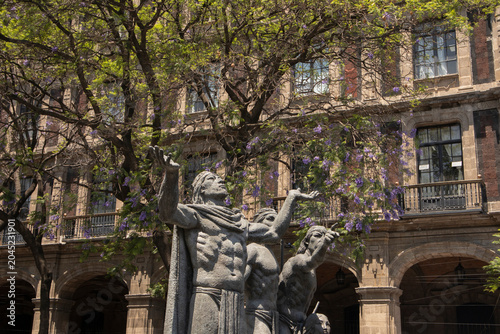A dramatic historic sculpture featuring human figures standing beneath blooming jacaranda trees in front of a grand colonial-style building.