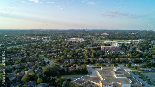 An expansive, high-angle aerial drone photograph looking across a green residential Oakville neighborhood with a public school in the foreground toward the distant Toronto and Mississauga skylines.