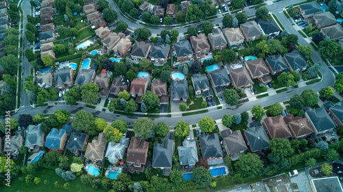 An expansive high-angle drone photograph looking straight down at a winding residential street that curves through an upscale suburban neighborhood featuring mature trees and backyard pools.