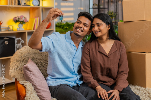 Indian couple man and woman sitting on home sofa show keys and smile with joy. Young family man and woman sitting on home sofa celebrate moving housing rental or purchase with boxes and fresh plans.
