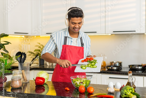 Indian man listening to music while mixing vegetable salad in home kitchen for healthy meal prep. Arabian guy smiling with headphones and tossing fresh greens for clean eating routine and diet food.
