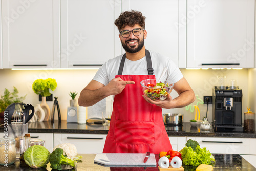 Arabian young man mixes fresh salad in bowl and speaks to camera recording cooking vlog in home kitchen. He explains keto diet and healthy eating routine while preparing vegetables for dinner at home.