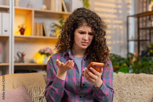 Young woman on home sofa reads bad news on smartphone then freezes in shock with hand on head from failure. Adult girl on home sofa shows stress disbelief defeat frustration during sad message check.