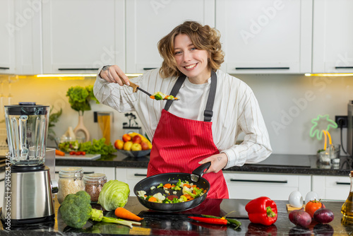 Young woman cooks vegetables in pan in kitchen for clean eating and healthy diet meal prep at home. Adult girl smiles while making fresh low calorie food for wellness weight loss and daily nutrition.