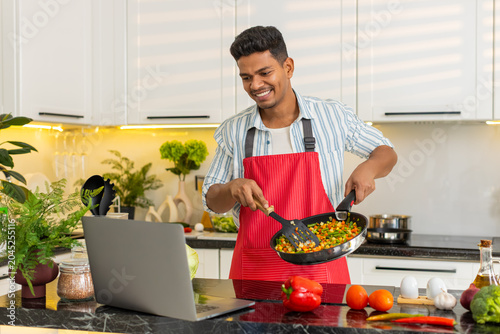 Indian man frying vegetables in pan while checking laptop for meal prep in home kitchen. Arabian guy smiling during diet routine with fresh food and simple lunch recipe for clean eating and nutrition.