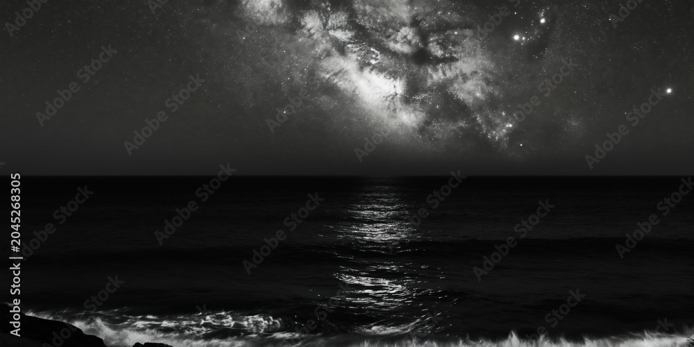 Obraz premium Black and white ocean expanse under starry sky. Dark sea water reflects faint moonlight. Gentle waves wash ashore on rocky coast at deep night.