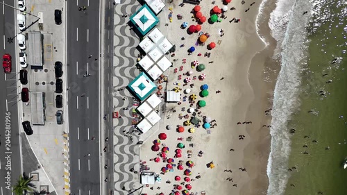 Top down aerial drone footage tracking along Copacabana Beach, following a vehicle beside the iconic wave-pattern promenade, colorful beach umbrellas, sandy shoreline and ocean waters in Rio de Janeir