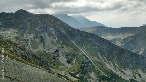 Dramatic mountain landscape of the Tatra Mountains in Poland under a cloudy sky with sharp rocky peaks, green slopes, and deep alpine valleys.
