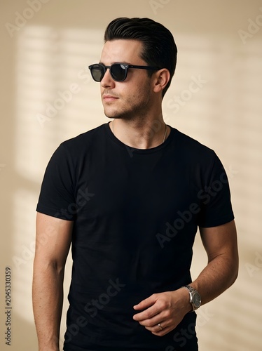 Handsome young man wearing blank black t-shirt and sunglasses against beige wall with window shadow