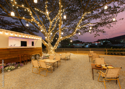 An inviting evening view of an outdoor camping-style cafe or event lawn, featuring wooden tables, folding canvas chairs on gravel, warm light bulb string garlands.