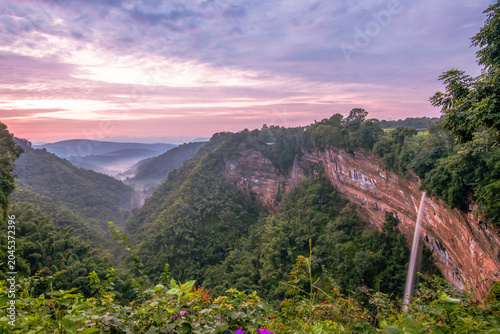 Tableau sur toile majestic waterfall flows down a red rock cliffside surrounded by a lush green forest valley covered in light mist under a beautiful purple pink sunrise sky