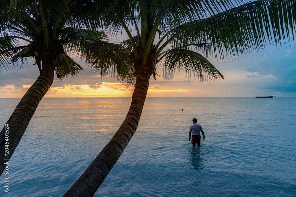 Fototapeta premium A man wades into the ocean at sunset in Koh Kood, Thailand, enjoying the tranquil evening waters. A cargo ship is visible on the horizon.